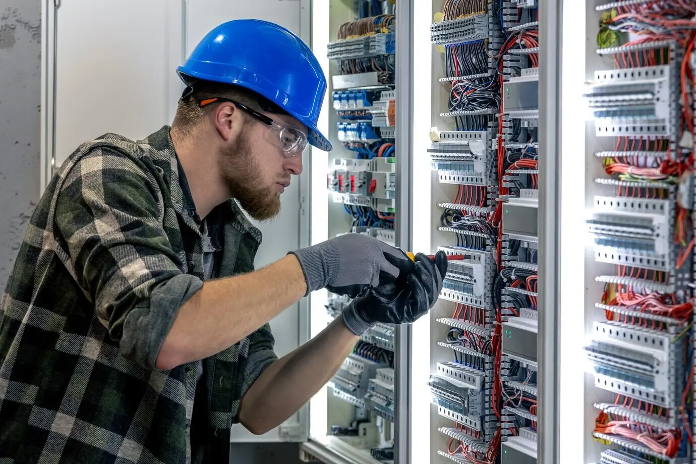An electrician inspects the switchboard wiring, checking it with a screwdriver in hand.