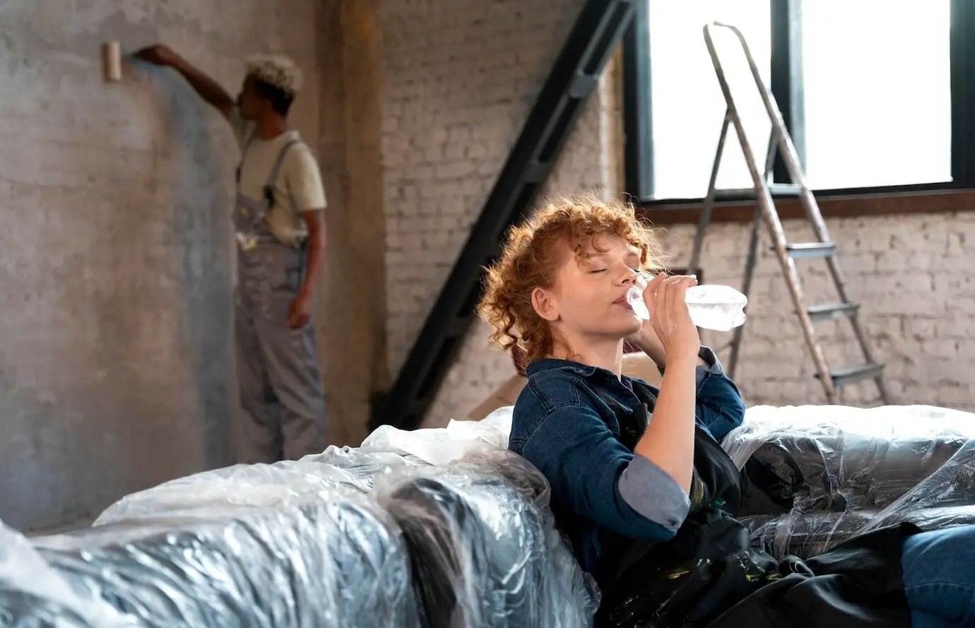 A woman drinks water as a man paints the wall of their new home.